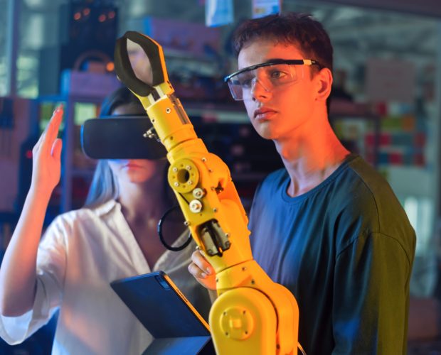 Teens doing experiments in robotics in a laboratory. Boy in protective glasses looking at a robot controlled by a girl hand. Red and blue illumination